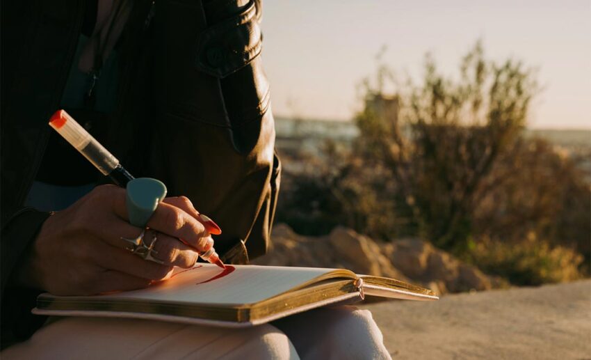 A woman sitting outdoors during a golden sunset, using a pen to write in an open notebook while surrounded by nature.