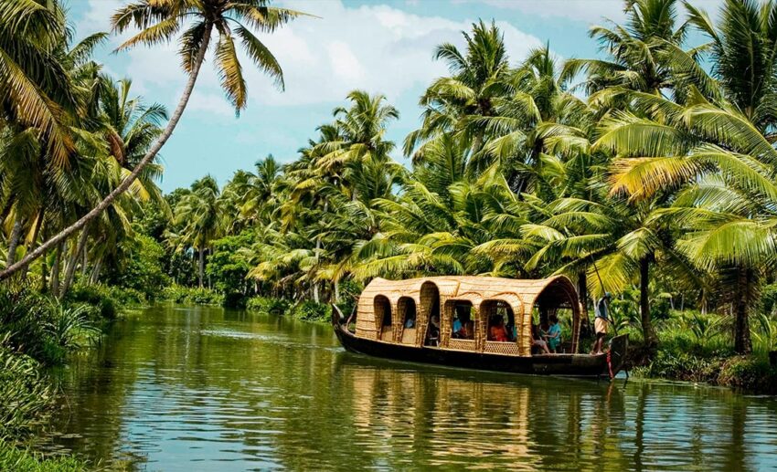 A traditional brown wooden boat on the calm backwaters of Kerala, India, surrounded by dense green palm trees.