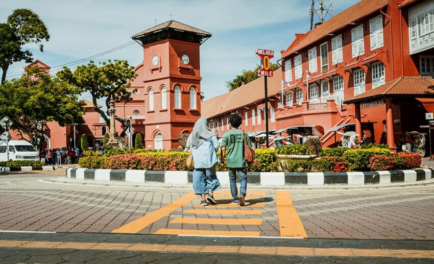 A man and a woman in a headscarf walking across the road to explore the historic red buildings of the Dutch Square in Melaka, Malaysia.