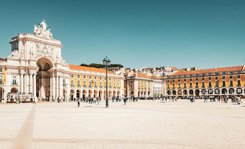 A wide shot of the historic Terreiro do Paço plaza in Lisbon, Portugal, with people walking around the grand yellow buildings during the daytime.