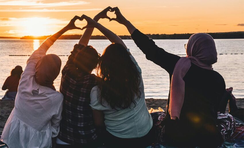 Four women sitting on a beach with their backs to the camera, silhouetted against a golden-hour sunset while forming heart shapes with their hands.
