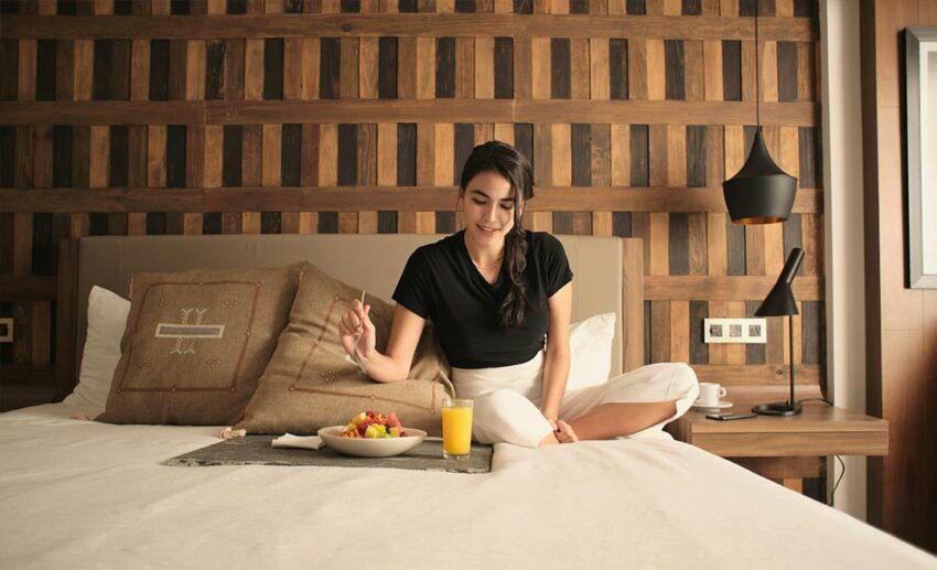 A woman sitting comfortably on a hotel bed with a tray of room-service breakfast in front of her in a bright, modern room.