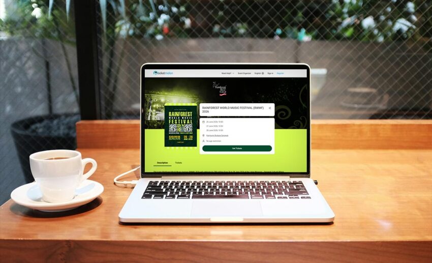 A laptop showing the official Rainforest World Music Festival ticket booking website on the screen, placed on a desk next to a coffee mug.