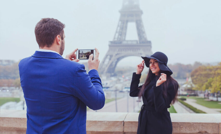 A man taking a photo of a woman with his smartphone as they stand as tourists near the Eiffel Tower in Paris.