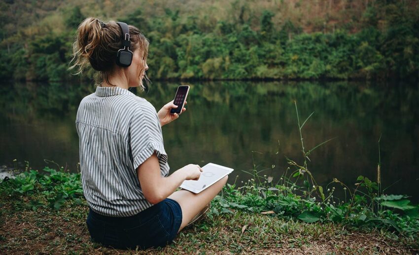 A woman sitting alone in a peaceful natural setting wearing headphones, holding a smartphone and a digital tablet to record or listen to ambient sounds.