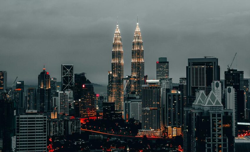 The Kuala Lumpur city skyline at dusk, featuring the illuminated Petronas Twin Towers against a deep blue and orange twilight sky.
