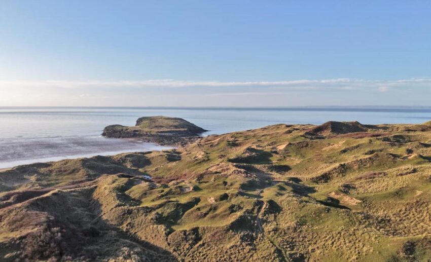 An aerial drone photograph showing the vast stretch of sandy beach and rolling green hills at Rhossili Bay.