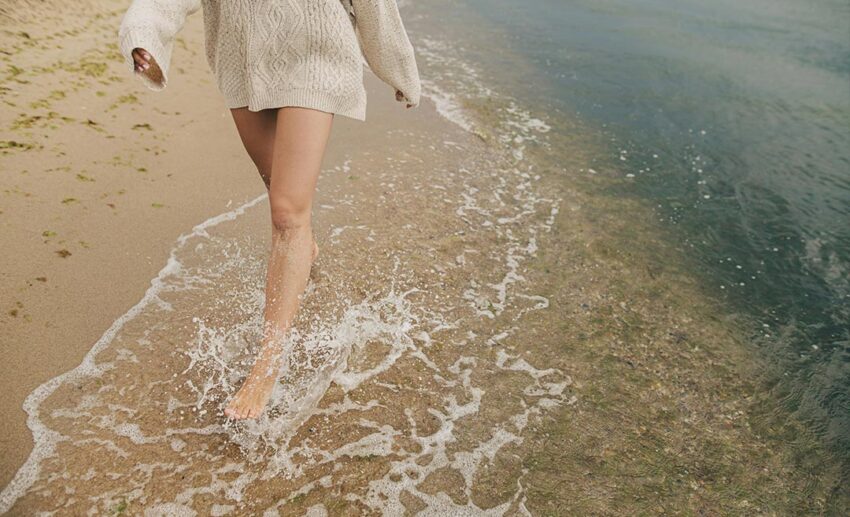 Cropped view of a woman’s feet running through cold sea waves on a sandy beach.
