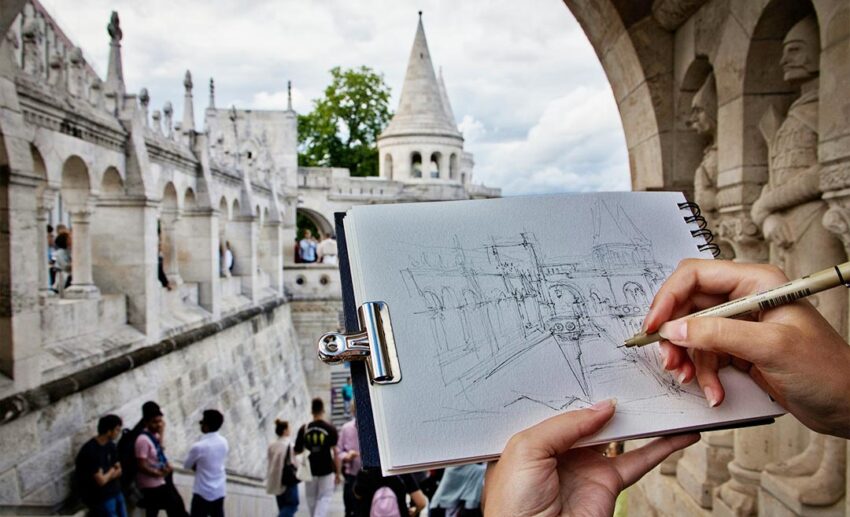 A close-up of a hand-drawn pencil sketch of Buda Castle in Budapest, Hungary, being held up in front of the actual landmark.