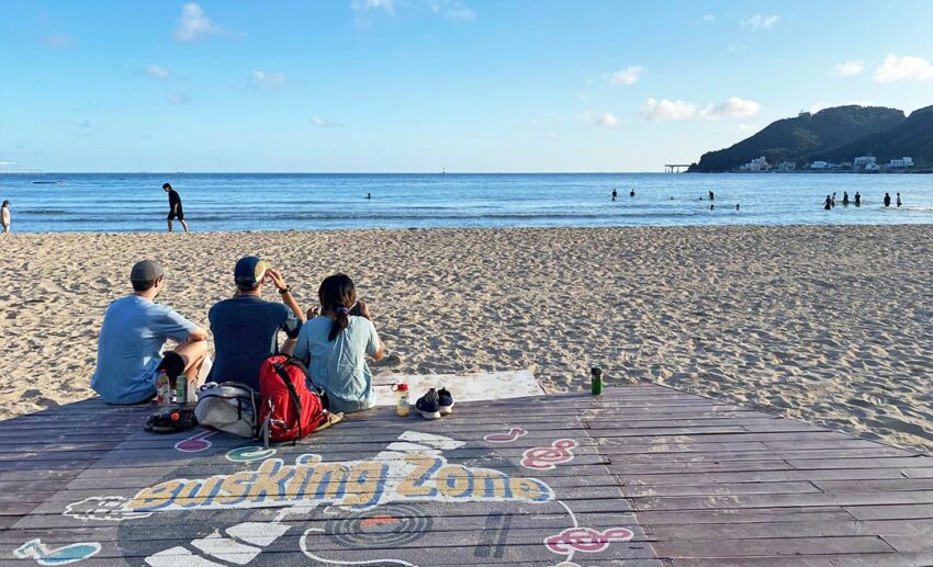 Three people sitting on a wooden busking deck at Songjeong Beach, looking out at the ocean in Busan.