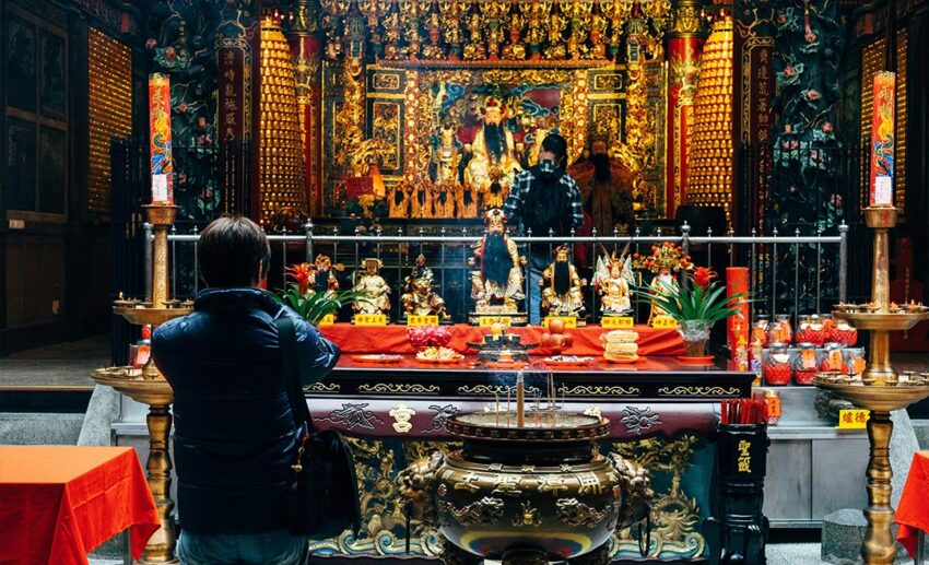 A woman standing at the entrance of a Buddhist temple, observing worshippers praying inside the serene, traditional structure.