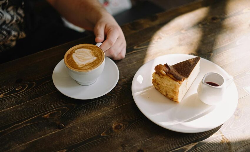 A white mug of coffee with latte art and a slice of cheesecake on a dark wooden cafe table.
