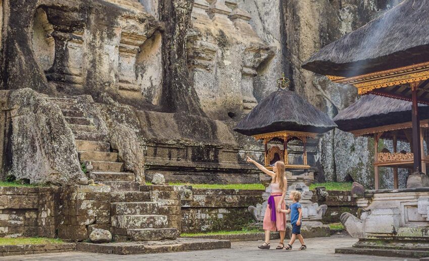A mother and son visiting the ancient stone-carved royal tombs of Gunung Kawi Temple in Ubud, Bali, surrounded by tropical nature.