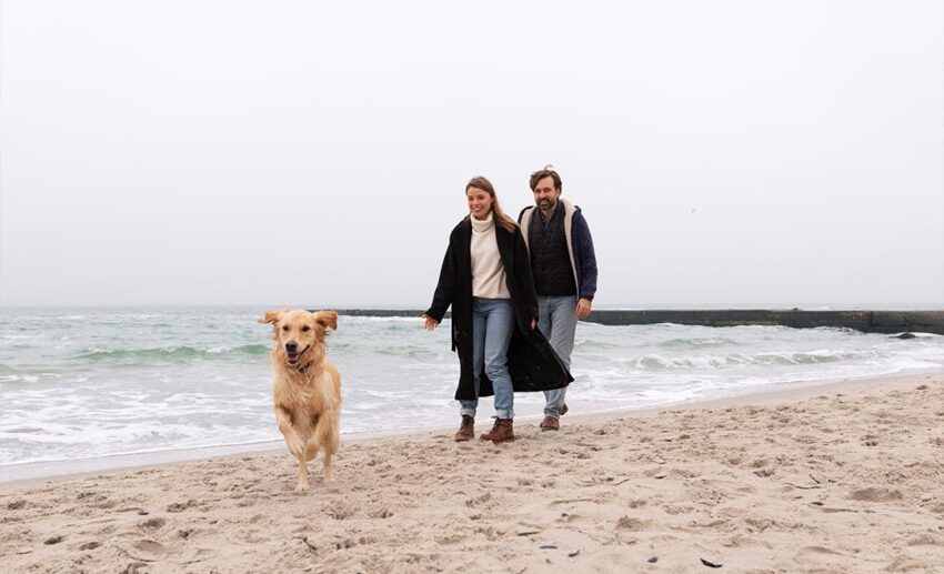 A local Norwegian couple smiling while walking along a scenic Bergen beach in winter, with their yellow Labrador Retriever running happily on the sand ahead of them.