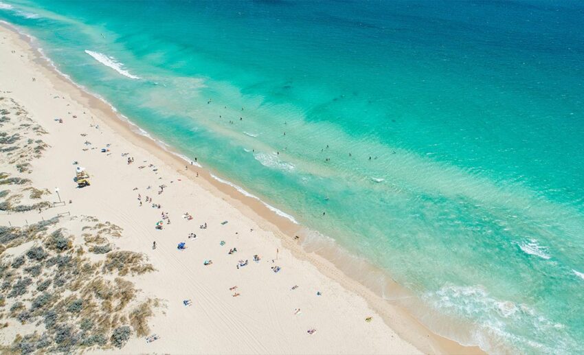 An aerial-style view of Scarborough Beach in Western Australia, showing turquoise waters, beige sand, and a few scattered swimmers and beachgoers during a quiet winter day.