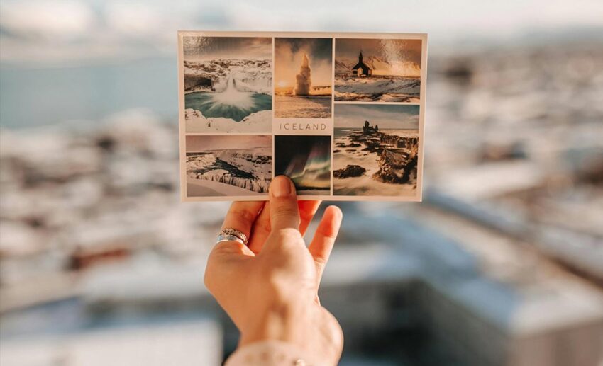 A close-up of a woman’s hand holding a scenic postcard of an Icelandic landscape featuring mountains and water.