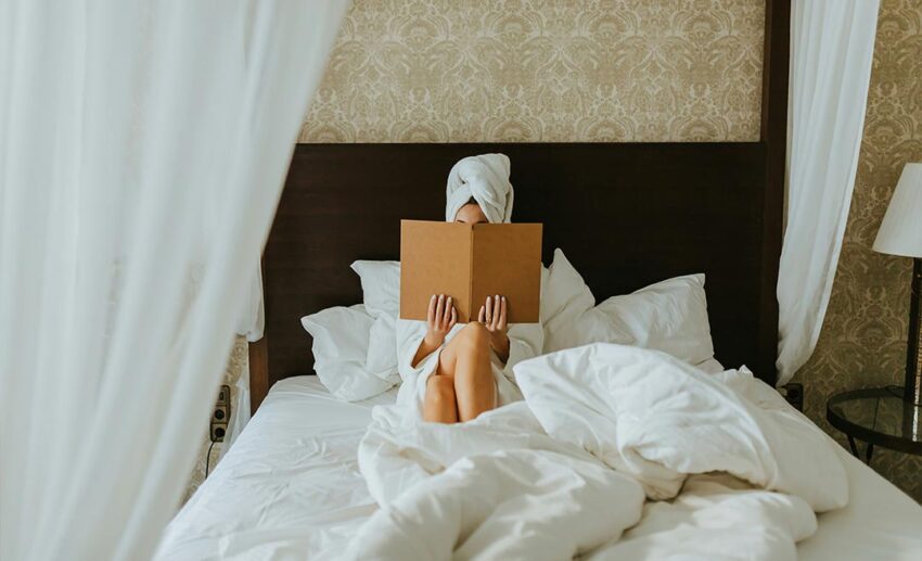 A woman relaxing on a hotel bed with a towel-wrapped head, reviewing an in-room menu in an air-conditioned room.