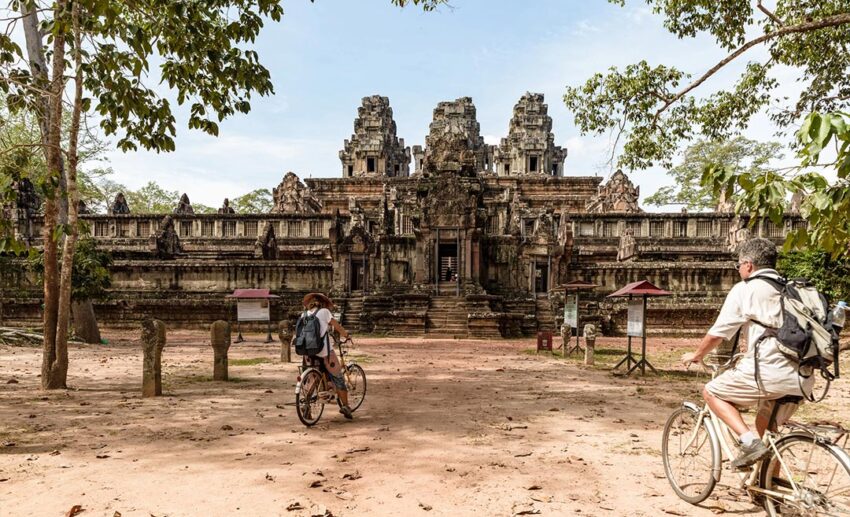 Two travellers cycling on a jungle path past the ancient Ta Keo temple ruins in Angkor, Cambodia, during the quiet rainy season.