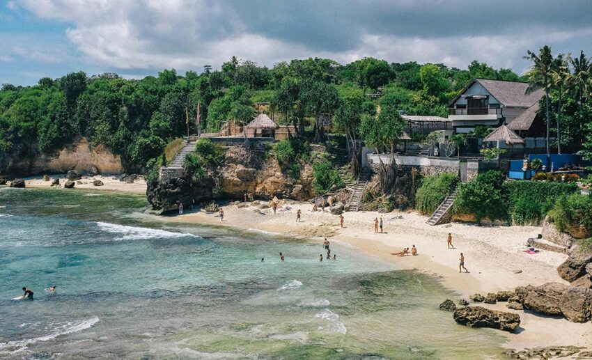 A distant shot of a secluded, rocky beach in Bali with people in the water and private luxury villas situated on the cliffs above the shoreline.