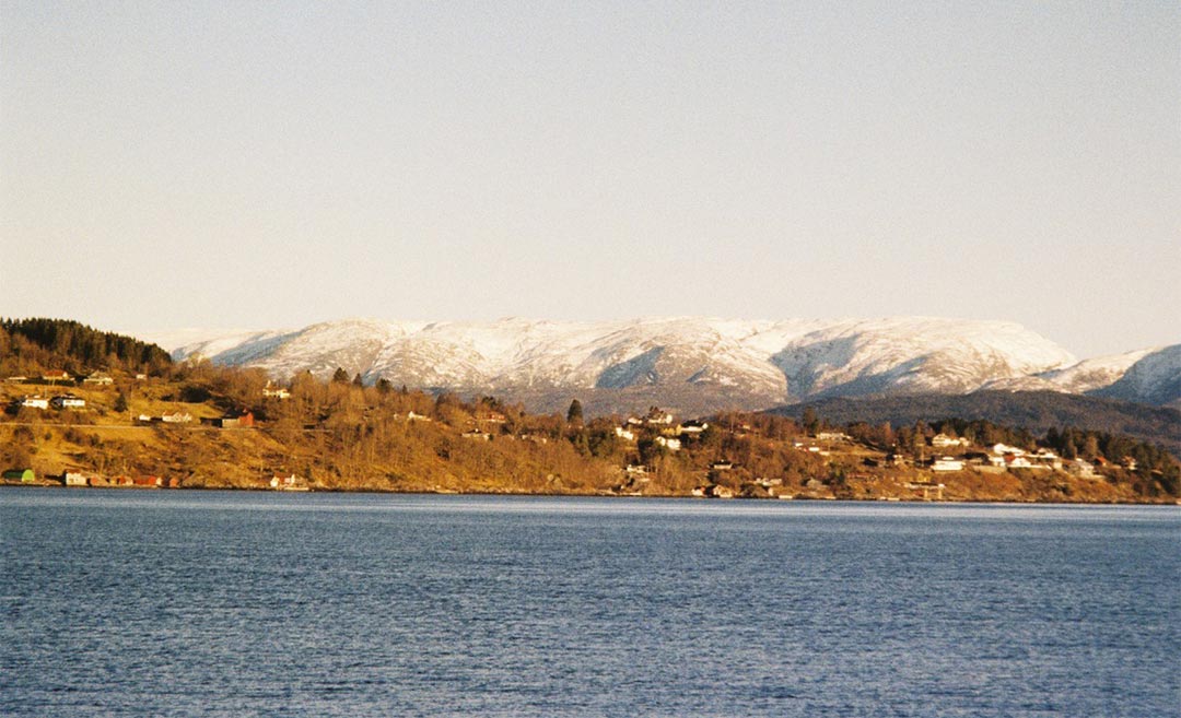A panoramic winter view of the Norwegian fjords in Bergen, featuring snow-capped mountains and sparsely placed traditional homes along the water’s edge.
