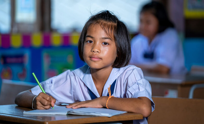 A young Malaysian elementary school girl sitting at a classroom desk, focused on her studies with a pencil and an open exercise book.