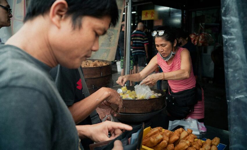 A woman stallkeeper in Southeast Asia picking siew mai from a rattan steamer into a plastic bag for a customer.