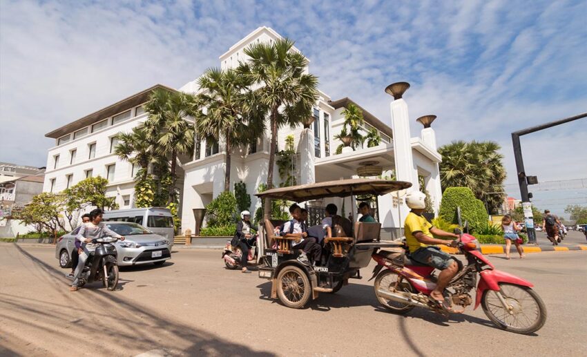 A traditional Cambodian tuk-tuk carrying passengers down a quiet, tree-lined road in Siem Reap.