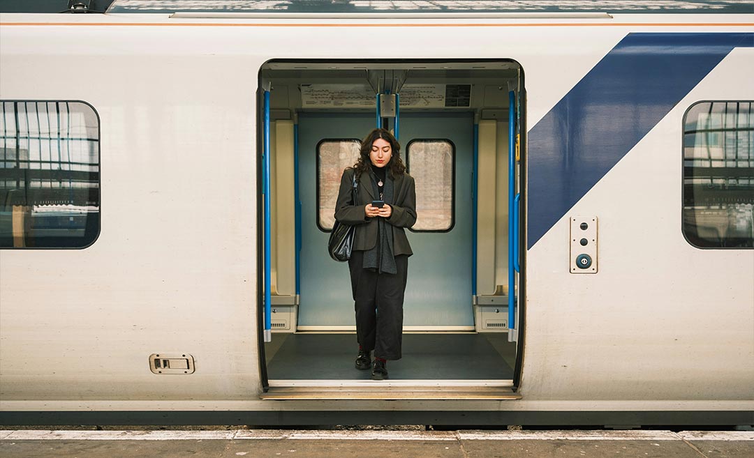 A solo female traveller standing in the open doorway of a stopped train, checking a map on her smartphone while looking out at the passing landscape.