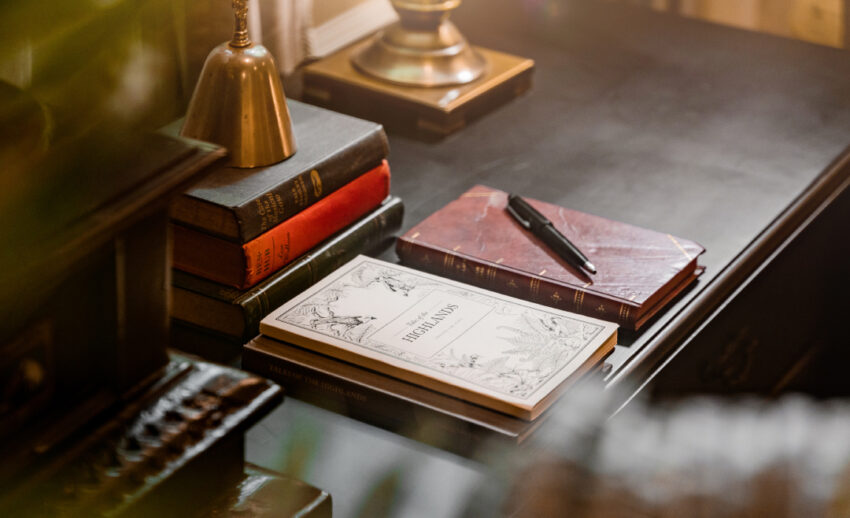 A close-up of a vintage-style dark wood desk featuring a stack of books, a classic desk lamp, and a fountain pen, symbolising the art of expressive writing.