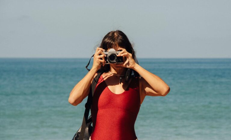 A woman holding a DSLR camera to her eye, about to take a photograph with the blue ocean in the background.