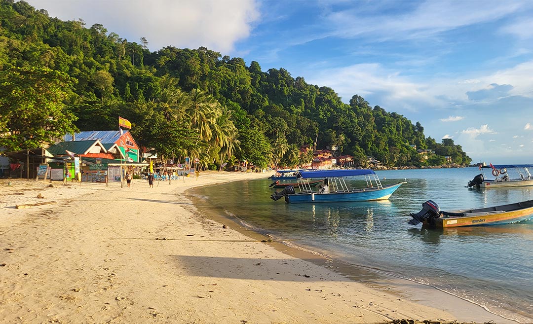 A daytime beach shot of Perhentian Kecil in Malaysia, featuring wooden boats anchored in shallow turquoise water near a colourful wooden building.