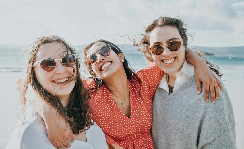 A supportive group of women standing together on a beach at sunset, with their arms around each other, celebrating a successful women's group trip.