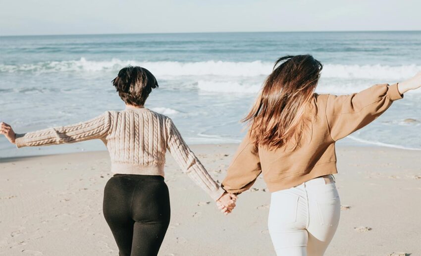 Two women walking hand-in-hand on a serene beach, embodying flexibility and calm during a group holiday.