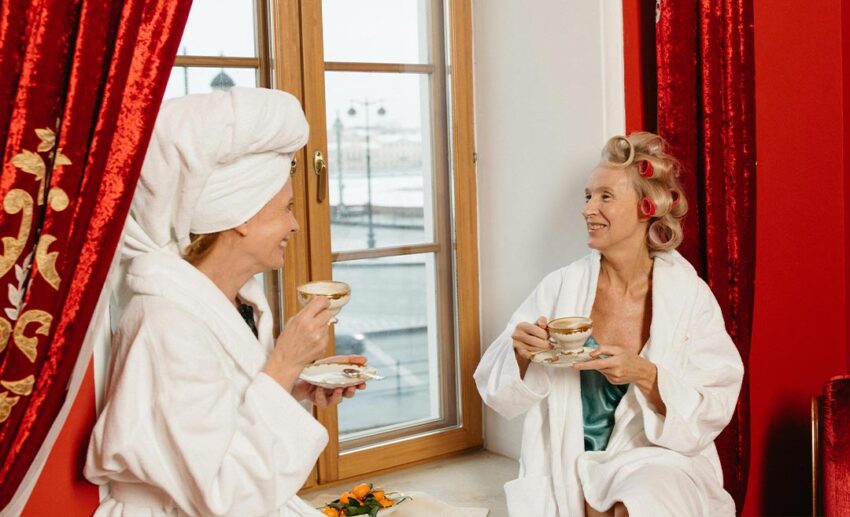 Two women in bathrobes sitting by a window and enjoying coffee during a spa retreat.