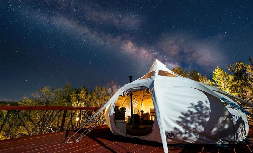 A luxury glamping tent illuminated from within against a dark, starry night sky in a remote nature location.