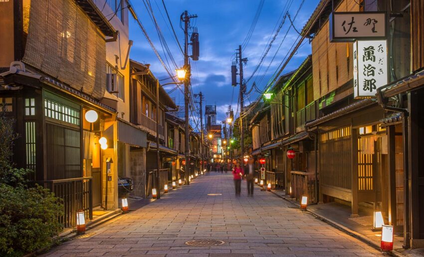 A view of Shinbashi-dori Street in Gion, Kyoto, at sunset, with traditional buildings and store lights beginning to glow.