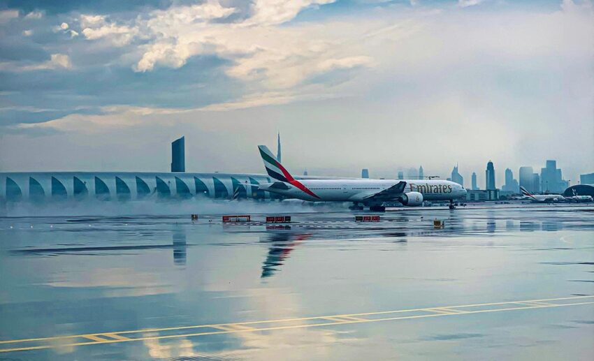 An Emirates airplane on a wet runway reflecting a cloudy sky, with a city skyline in the background, representing the fragile recovery of flight routes.