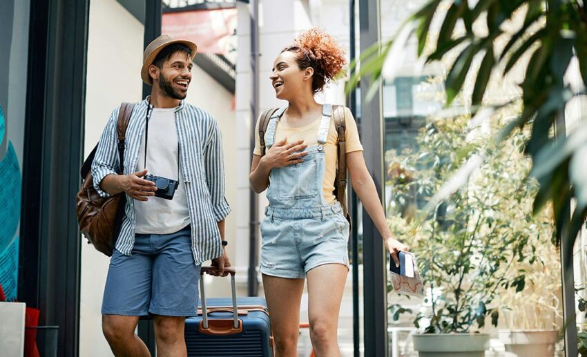 A smiling young couple chatting while walking into a hotel lobby, portraying travellers who have successfully adapted their arrival plans to suit later check-in schedules.