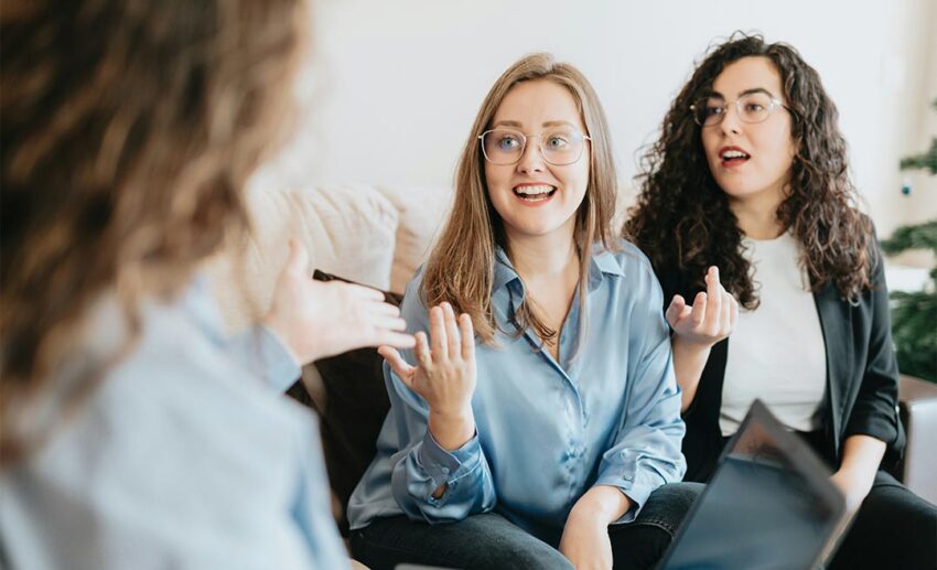 Three women sitting comfortably on a couch having an inclusive conversation about their upcoming group holiday plans.