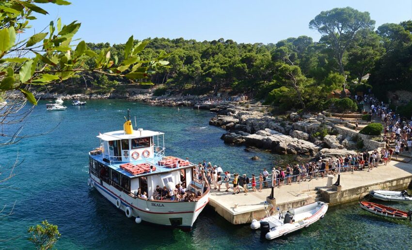 A group of travellers standing on a stone dock next to a boat with the historic coastal landscape of Dubrovnik, Croatia, in the background.