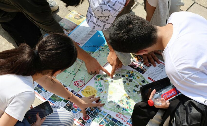 A group of young adult travellers standing outdoors, excitedly examining a city map and pointing at specific locations for their itinerary.