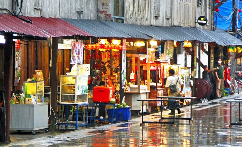 The wet, reflective streets of the Siniawan night market in Malaysia just after a monsoonal downpour, with vendors seen in the background beginning to reset their stalls as the rain clears.