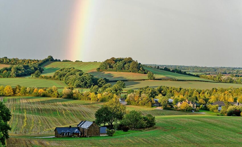 A scenic view of a small historic town in the Cotswolds, United Kingdom, with a rainbow arching over stone buildings during the autumn season.