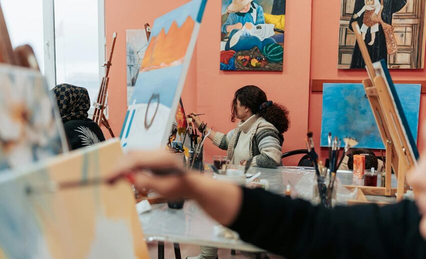 A woman focused on painting a colourful canvas in a light-filled art studio setting.