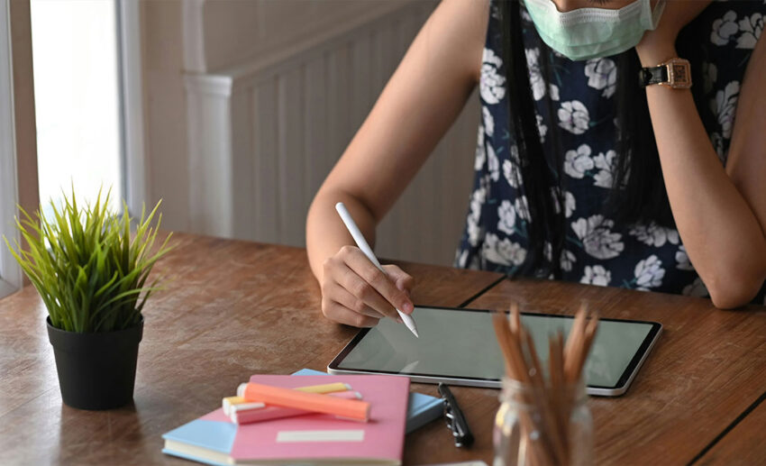 A woman wearing a blue medical face mask while focused on drawing on a computer tablet with a stylus pen in a quiet indoor setting.