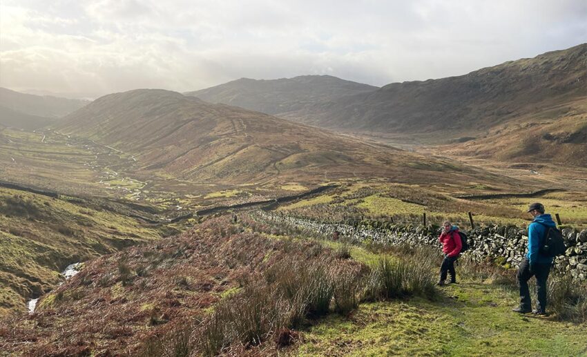 The writer’s travel companions walking along a lush, green countryside path in the Lake District during a summer hiking trip.