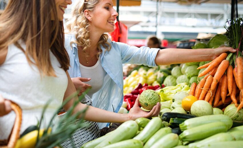 A young woman smiling while shopping for fresh fruits and vegetables at a vibrant outdoor farmers' market.