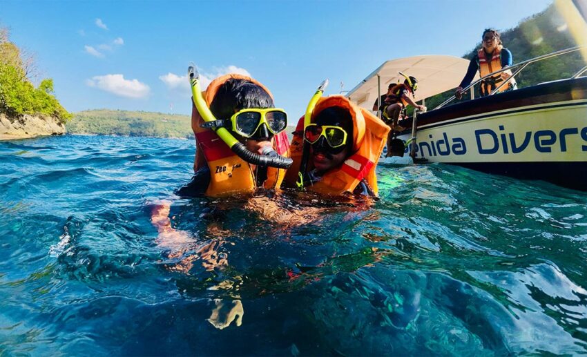 Two children wearing life jackets and snorkel gear swimming in the clear blue waters near Nusa Penida, Bali, during a family holiday.