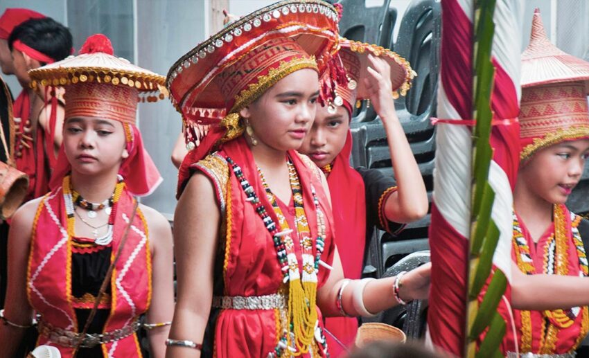 Young Dayak girls dressed in intricate red, black, and gold traditional clothing and headgear during a harvest festival celebration in East Malaysia.