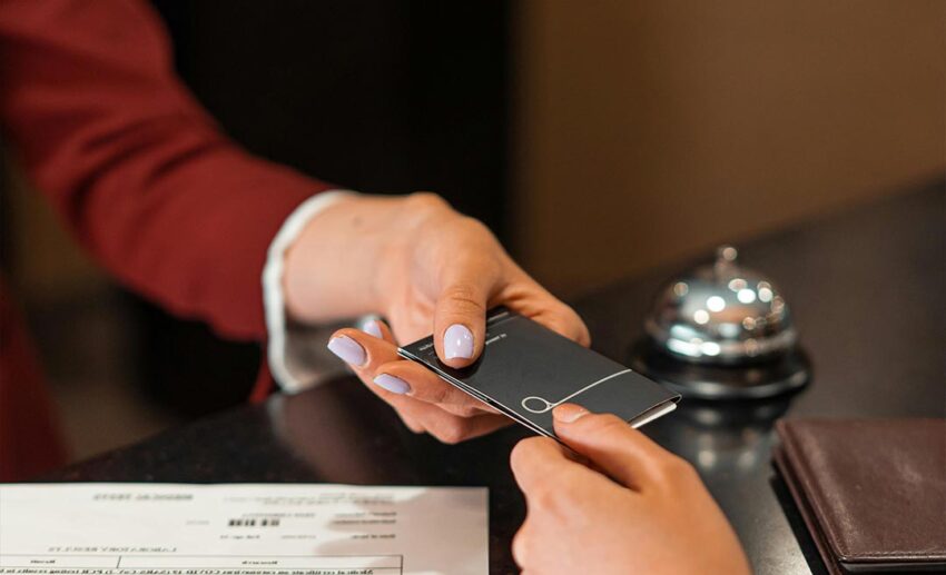 A close-up of a hotel receptionist handing a digital key card to a guest across a wooden counter, representing the delay in room readiness due to operational shifts.